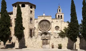 Kamienne mury klasztoru w Sant Cugat del Vallès, ozdobione rozetą i wieżą z dzwonami, otoczone są strzelistymi cyprysami.
