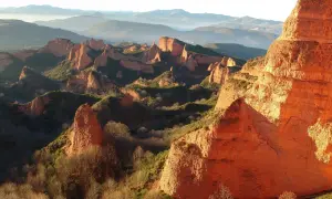 Imponujące, pomarańczowe formacje skalne Las Médulas w promieniach zachodzącego słońca, otoczone zielenią i mglistymi górami w tle.