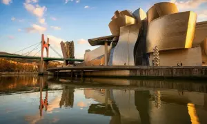 Muzeum Guggenheima w Hiszpanii, Bilbao, lśni w słońcu, odbijając się w rzece Nervión. Mosty i miasto tworzą malowniczy krajobraz.