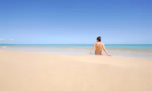 Kobieta siedzi na piaszczystej plaży, patrząc na turkusowe morze. To idealne miejsce na plaży dla nudystów, gdzie można poczuć wolność.