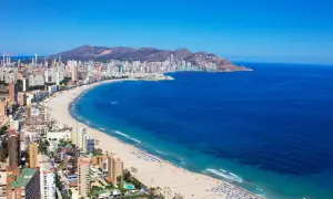 Słoneczna pogoda na Costa Blanca: szeroka plaża z turystami, turkusowe morze i miasto z drapaczami chmur w tle.