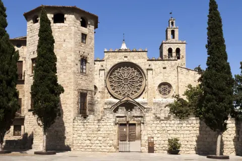 Kamienne mury klasztoru w Sant Cugat del Vallès, ozdobione rozetą i wieżą z dzwonami, otoczone są strzelistymi cyprysami.