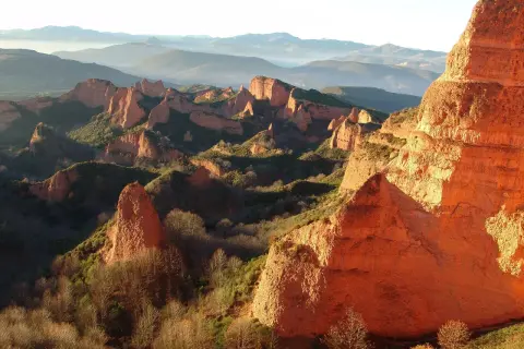 Imponujące, pomarańczowe formacje skalne Las Médulas w promieniach zachodzącego słońca, otoczone zielenią i mglistymi górami w tle.