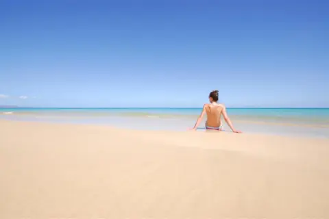 Kobieta siedzi na piaszczystej plaży, patrząc na turkusowe morze. To idealne miejsce na plaży dla nudystów, gdzie można poczuć wolność.