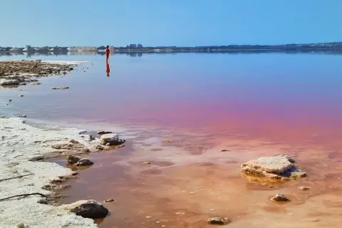 Osoba w czerwonym stroju stoi w różowym jeziorze w Hiszpanii. W tle widoczne są hałdy soli i zabudowania.