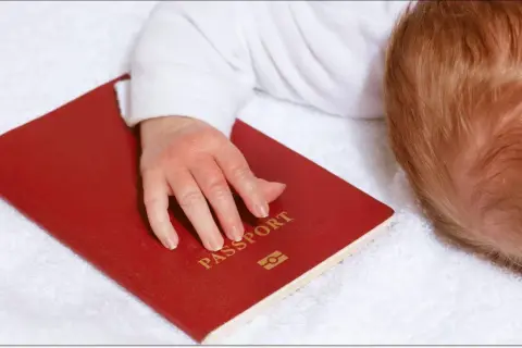 Dziecko z rudymi włosami śpi na białym ręczniku, jego dłoń spoczywa na czerwonym paszporcie. Czy do Hiszpanii trzeba paszport?