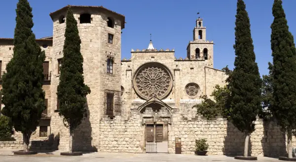 Kamienne mury klasztoru w Sant Cugat del Vallès, ozdobione rozetą i wieżą z dzwonami, otoczone są strzelistymi cyprysami.