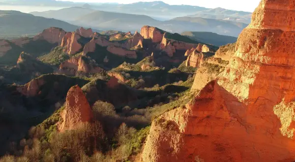 Imponujące, pomarańczowe formacje skalne Las Médulas w promieniach zachodzącego słońca, otoczone zielenią i mglistymi górami w tle.