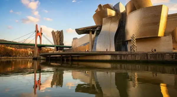 Muzeum Guggenheima w Hiszpanii, Bilbao, lśni w słońcu, odbijając się w rzece Nervión. Mosty i miasto tworzą malowniczy krajobraz.