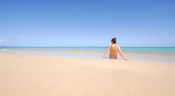 Kobieta siedzi na piaszczystej plaży, patrząc na turkusowe morze. To idealne miejsce na plaży dla nudystów, gdzie można poczuć wolność.