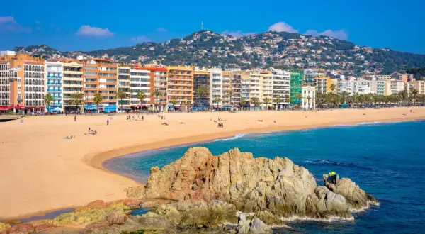 Słoneczna plaża w Lloret de Mar z kolorowymi budynkami w tle i skalistym wybrzeżem.