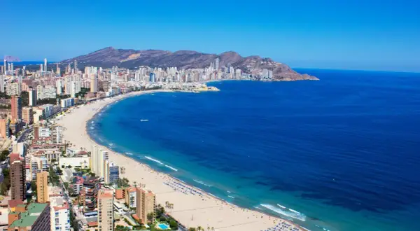 Słoneczna pogoda na Costa Blanca: szeroka plaża z turystami, turkusowe morze i miasto z drapaczami chmur w tle.