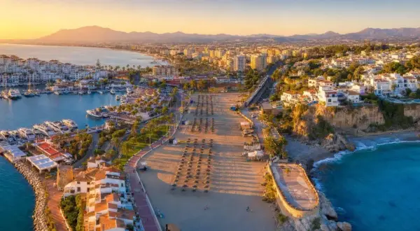 Złociste słońce oświetla port i pustą plażę w Nerja. Długi deptak z rzędami parasoli prowadzi do turkusowego morza.