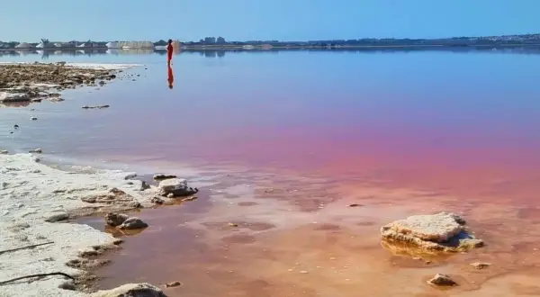 Osoba w czerwonym stroju stoi w różowym jeziorze w Hiszpanii. W tle widoczne są hałdy soli i zabudowania.