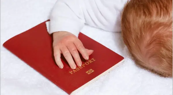 Dziecko z rudymi włosami śpi na białym ręczniku, jego dłoń spoczywa na czerwonym paszporcie. Czy do Hiszpanii trzeba paszport?