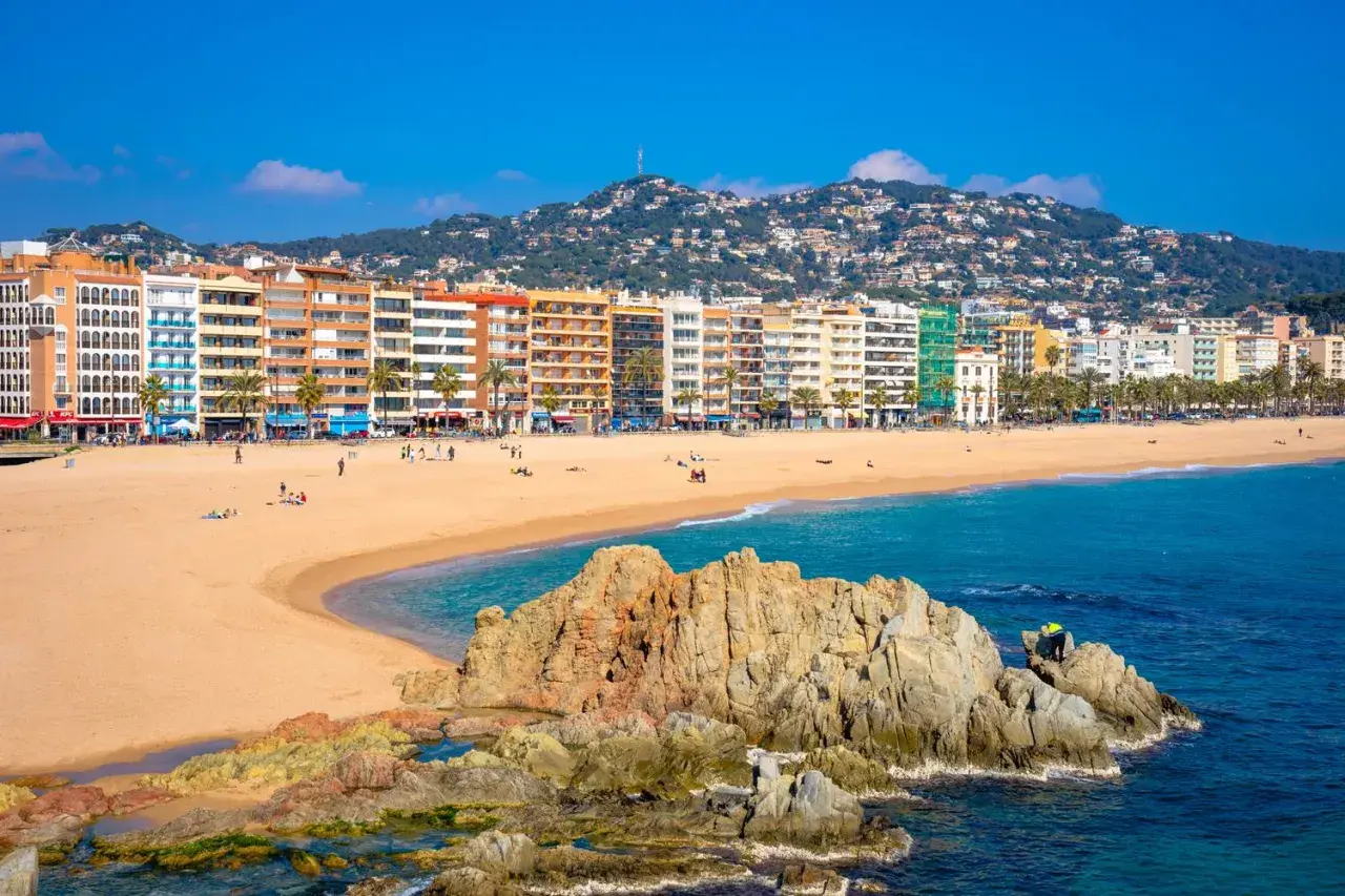 Słoneczna plaża w Lloret de Mar z kolorowymi budynkami w tle i skalistym wybrzeżem.