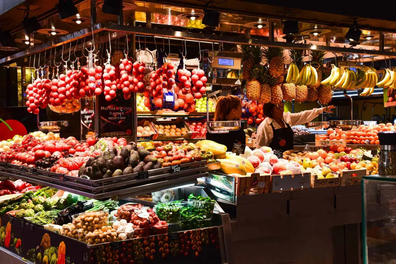 Kolorowe stoisko z owocami i warzywami na targu La Boqueria w Barcelonie. Wiszące pomidory, banany, ananasy, papaje, awokado i wiele innych świeżych produktów.