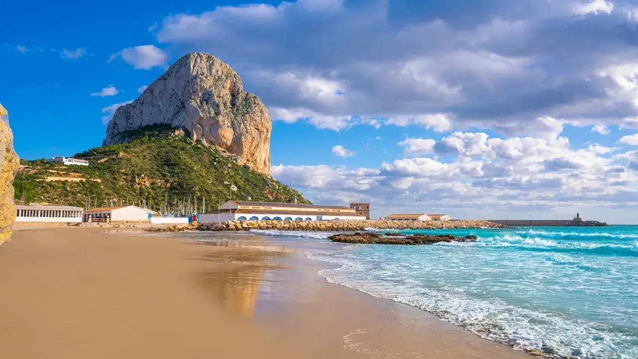 Piękna pogoda w Calpe: skalista góra Peñón de Ifach, plaża, turkusowe morze i białe budynki portu.