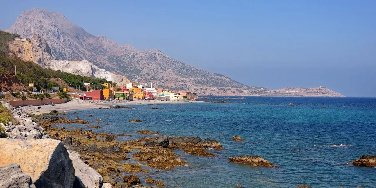 Kolorowe domy nad zatoką w Ceuta, Hiszpania, z górami w tle i skalistym wybrzeżem.