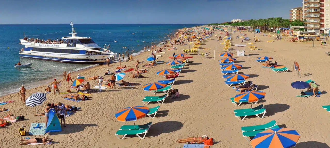 Słoneczna plaża w Santa Susanna, pełna ludzi opalających się i odpoczywających pod parasolami. Statek wycieczkowy