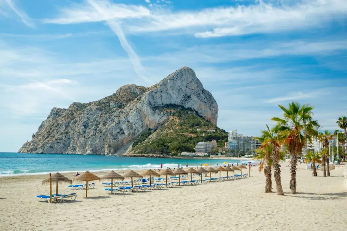 Piękny dzień na wybrzeżu Costa Blanca. Piaszczysta plaża z leżakami i parasolami, turkusowe morze i majestatyczna skała w tle.