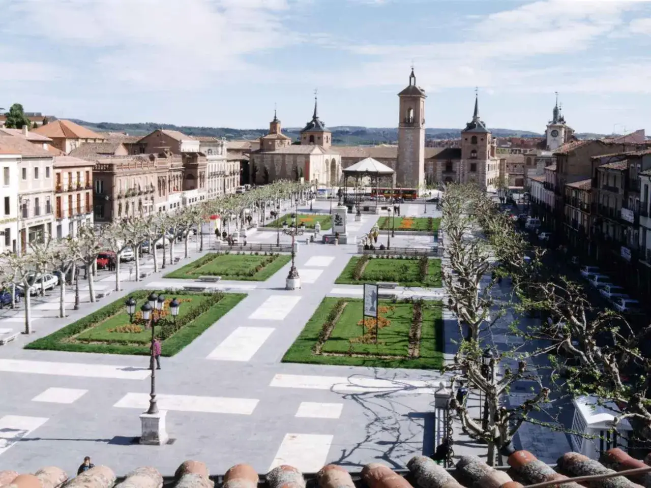 Plac w Alcalá de Henares z alejami drzew, trawnikami i zabytkowymi budynkami.