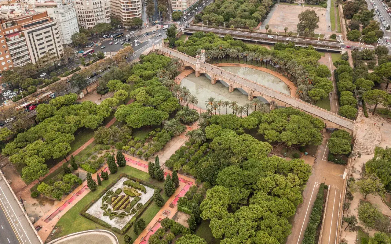 Widok z lotu ptaka na park Turia z kamiennym mostem nad wodą, otoczony bujną zielenią i budynkami w tle.