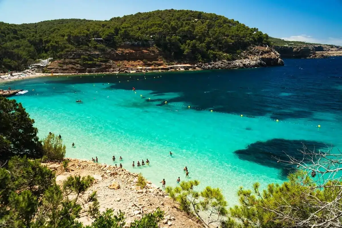Malownicza cala bassa z turkusową wodą, skalistymi brzegami i zielonymi wzgórzami. Ludzie kąpią się i relaksują na plaży.
