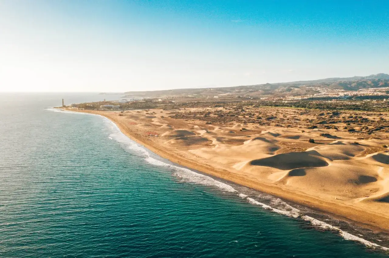 Z lotu ptaka widać turkusowe morze i piaszczyste wydmy Maspalomas. W oddali majaczy latarnia morska i zabudowania.