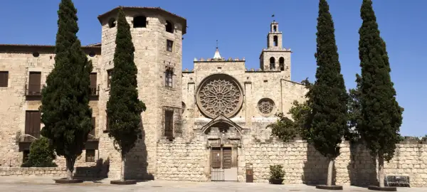Kamienne mury klasztoru w Sant Cugat del Vallès, ozdobione rozetą i wieżą z dzwonami, otoczone są strzelistymi cyprysami.