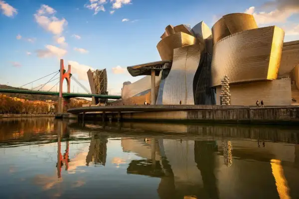 Muzeum Guggenheima w Hiszpanii, Bilbao, lśni w słońcu, odbijając się w rzece Nervión. Mosty i miasto tworzą malowniczy krajobraz.