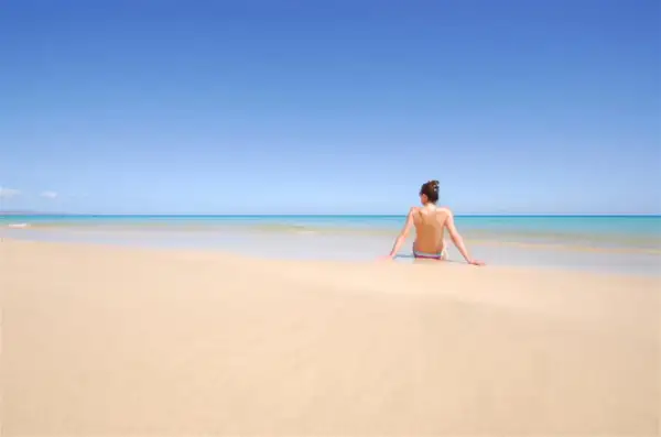 Kobieta siedzi na piaszczystej plaży, patrząc na turkusowe morze. To idealne miejsce na plaży dla nudystów, gdzie można poczuć wolność.