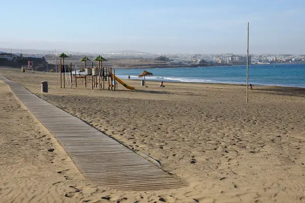 Plaża w stolicy Fuerteventury, z placem zabaw i drewnianą ścieżką. W tle widać ocean i zabudowania miasta.