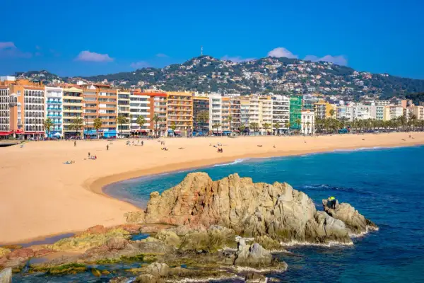 Słoneczna plaża w Lloret de Mar z kolorowymi budynkami w tle i skalistym wybrzeżem.