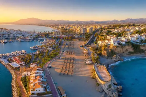 Złociste słońce oświetla port i pustą plażę w Nerja. Długi deptak z rzędami parasoli prowadzi do turkusowego morza.