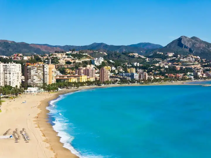 Malaga plaża z turkusową wodą i zabudowaniami na wzgórzach. Słoneczny dzień nad Morzem Śródziemnym.